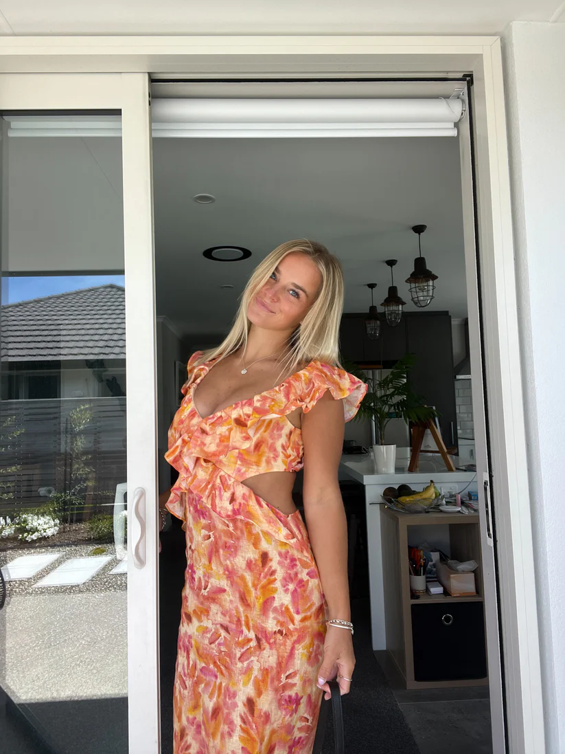 Smiling woman in a vibrant orange and pink floral cutout maxi dress with ruffled sleeves and a deep V-neckline.