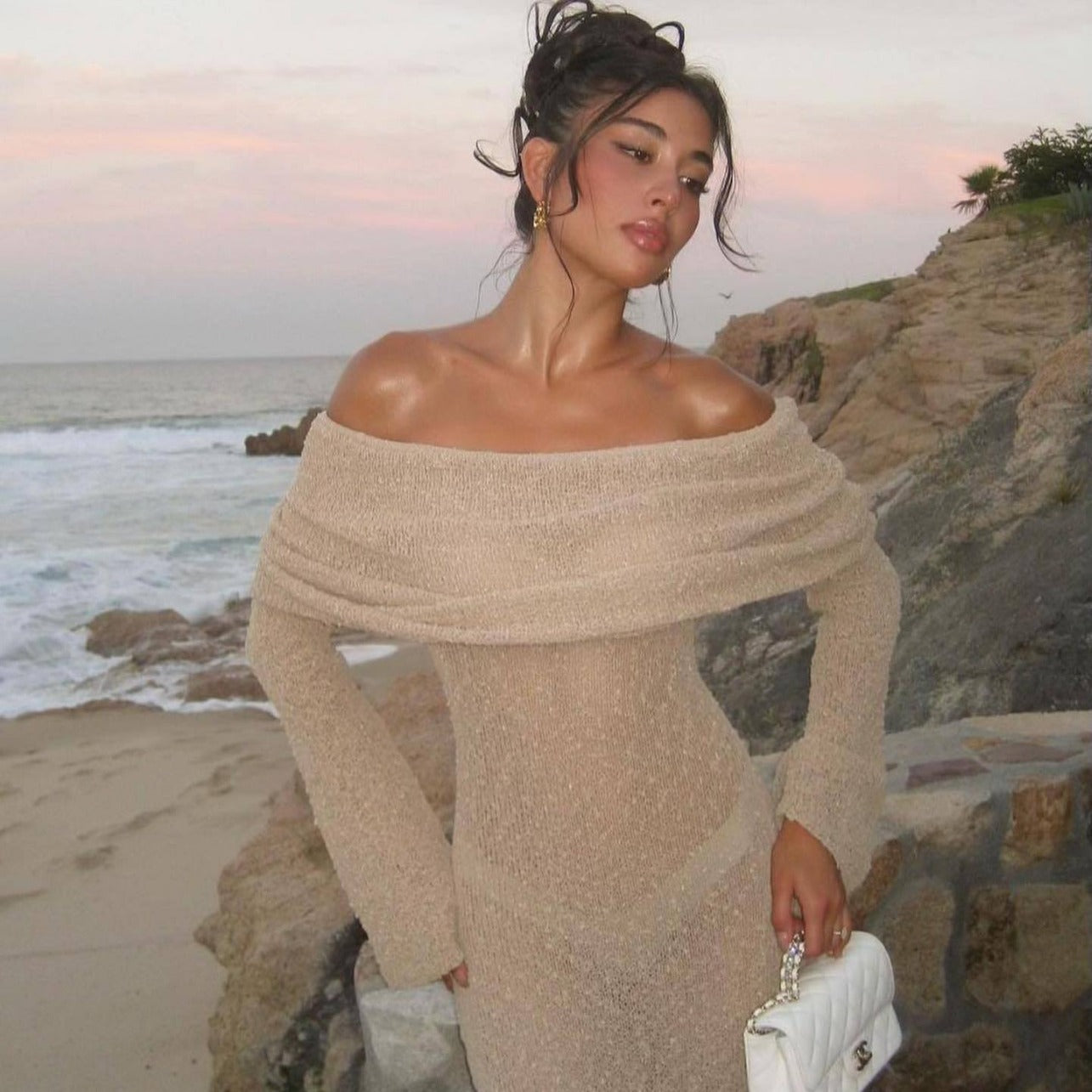 Beige Valencia Drape Maxi Dress with off-the-shoulder design, long sleeves, and a white quilted handbag, on a beach at sunset.