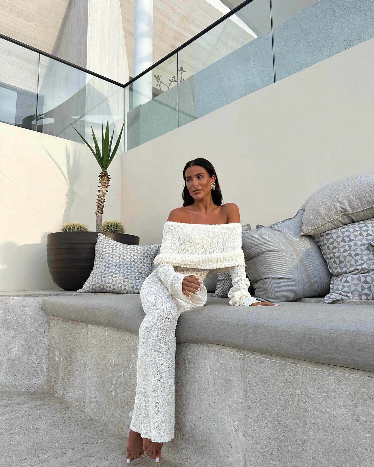 White Valencia Drape Maxi Dress with an off-the-shoulder neckline, long sleeves, and a form-fitting silhouette, worn while seated.