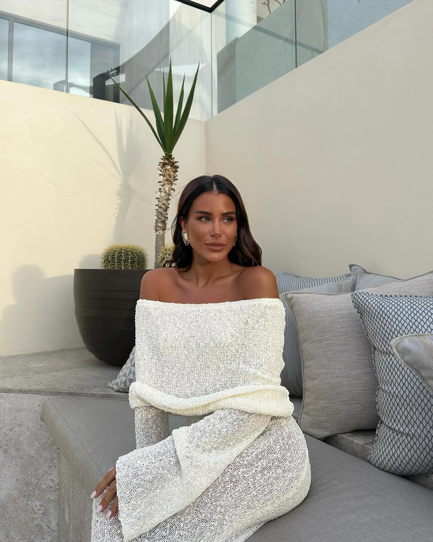 Close-up of white Valencia Drape Maxi Dress, showcasing the off-the-shoulder design and textured knit fabric while seated.