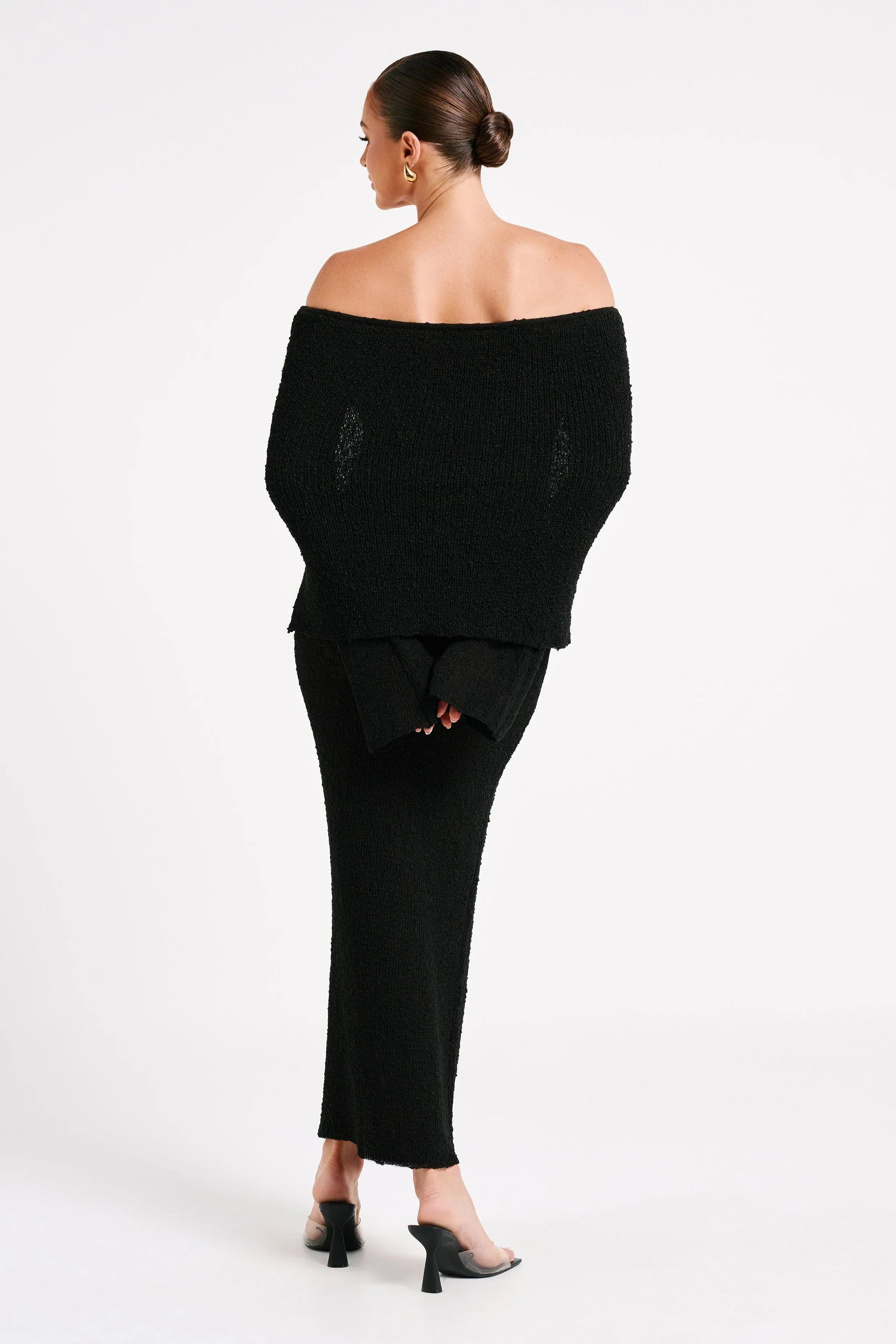 Back view of black Valencia Drape Maxi Dress, featuring an off-the-shoulder neckline and long sleeves, paired with dark heels.
