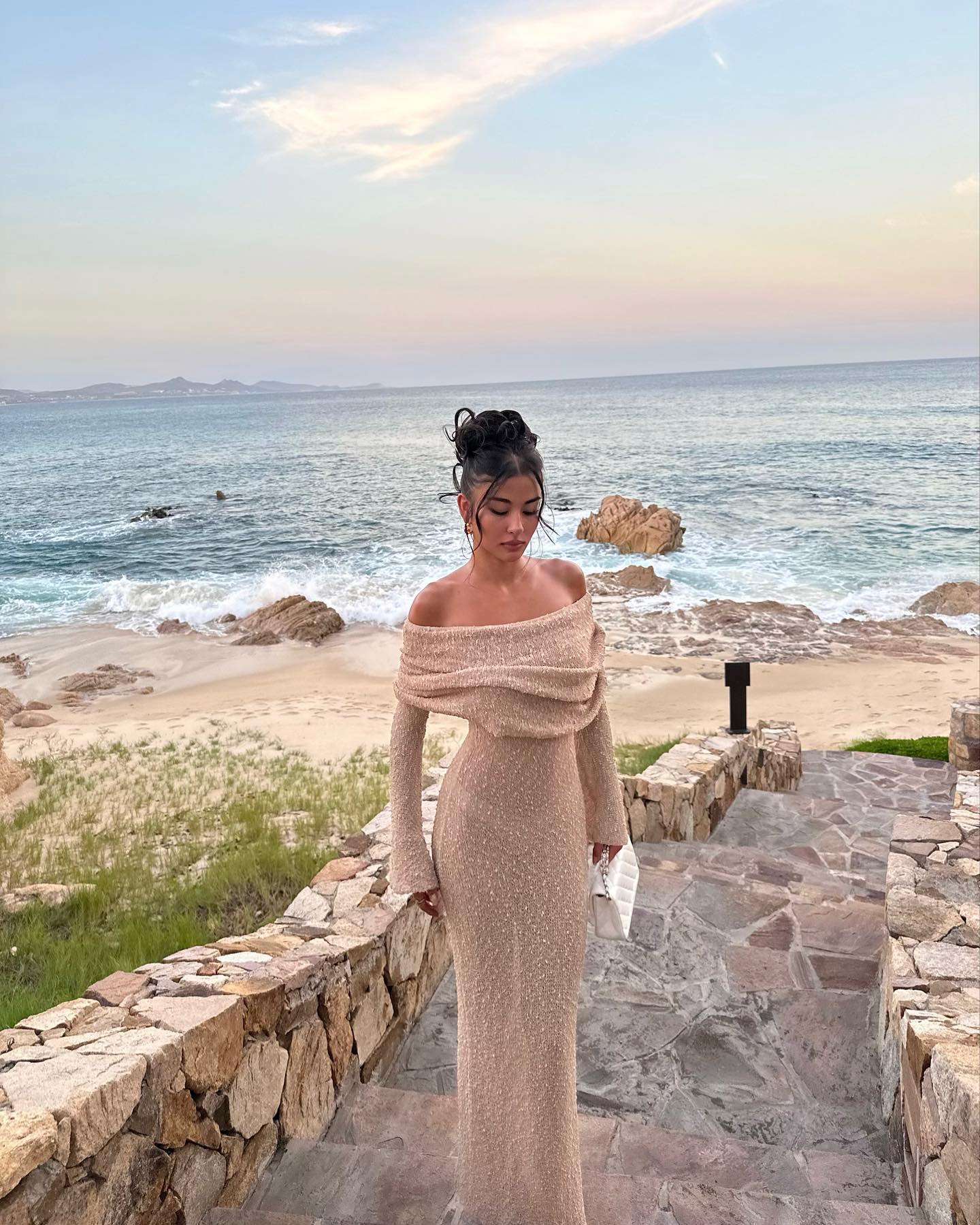 Beige Valencia Drape Maxi Dress with off-the-shoulder detail and long sleeves, worn on stone steps leading to a beach.
