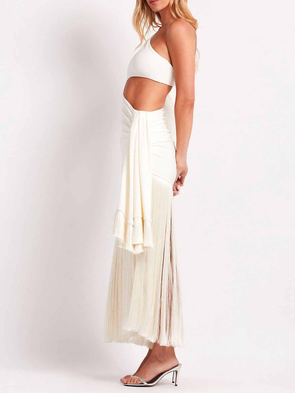 Side profile of a white one-shoulder maxi dress with a side cutout, long sleeve, and a flowing fringe skirt.