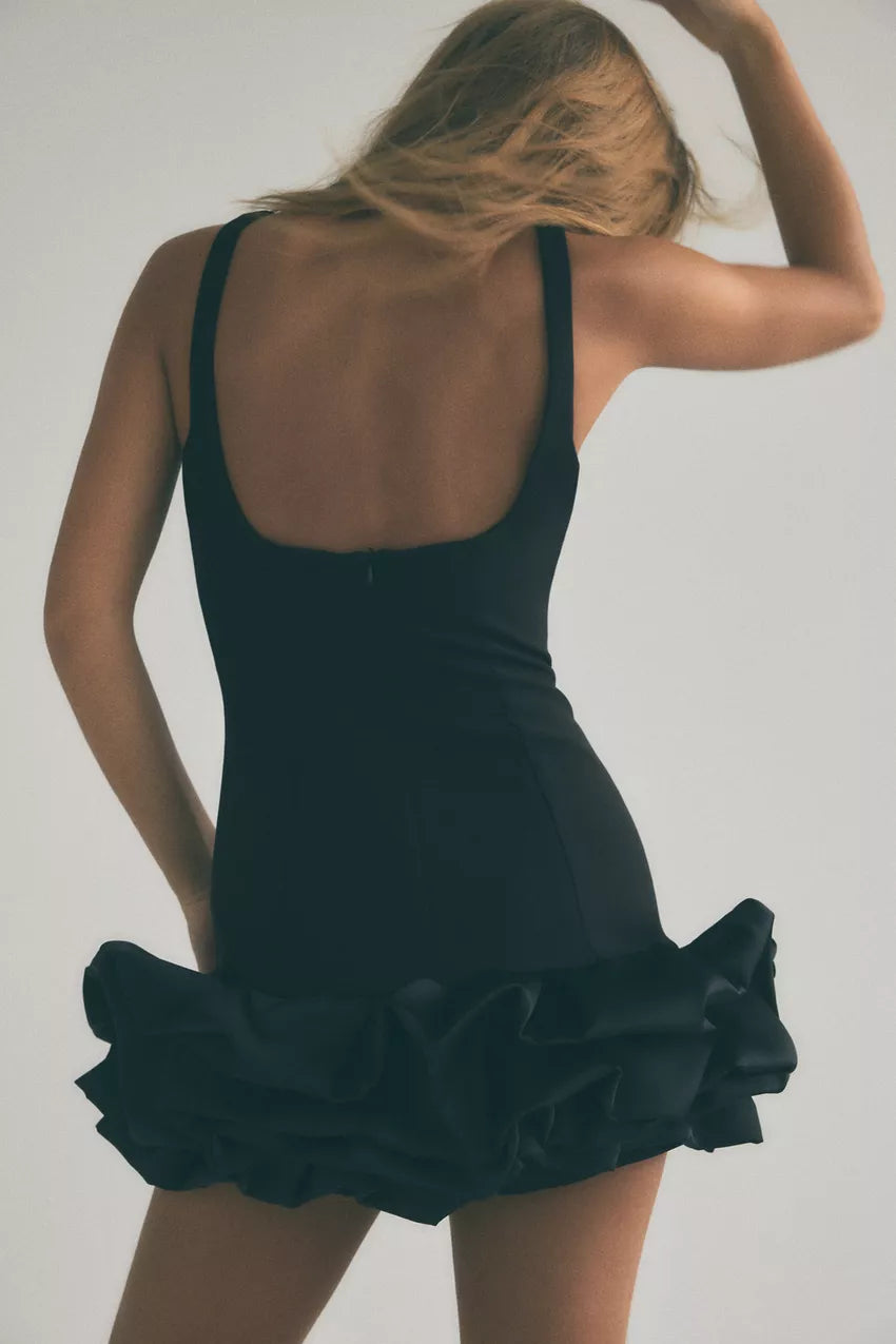 Back view of a model in a black Vienna Rosette Mini Dress, showcasing the scoop back and ruffled skirt.