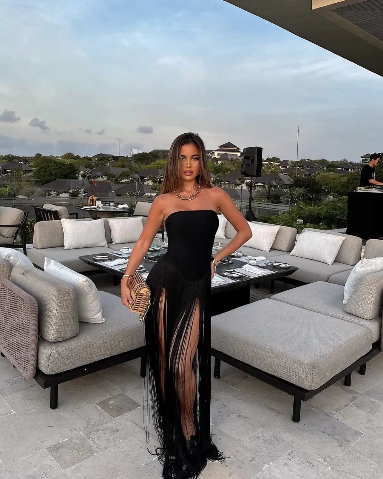 Full view of a black strapless violet fringe maxi dress with a high slit, accessorized with a gold chain necklace.