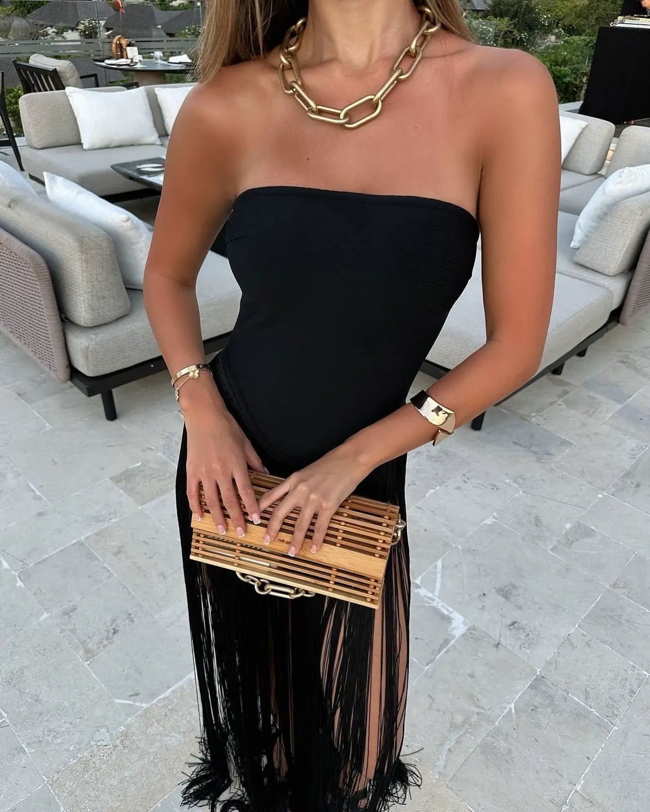 Close-up of the black strapless bodice and violet fringe skirt of a maxi dress, with a gold chain necklace and wooden clutch.