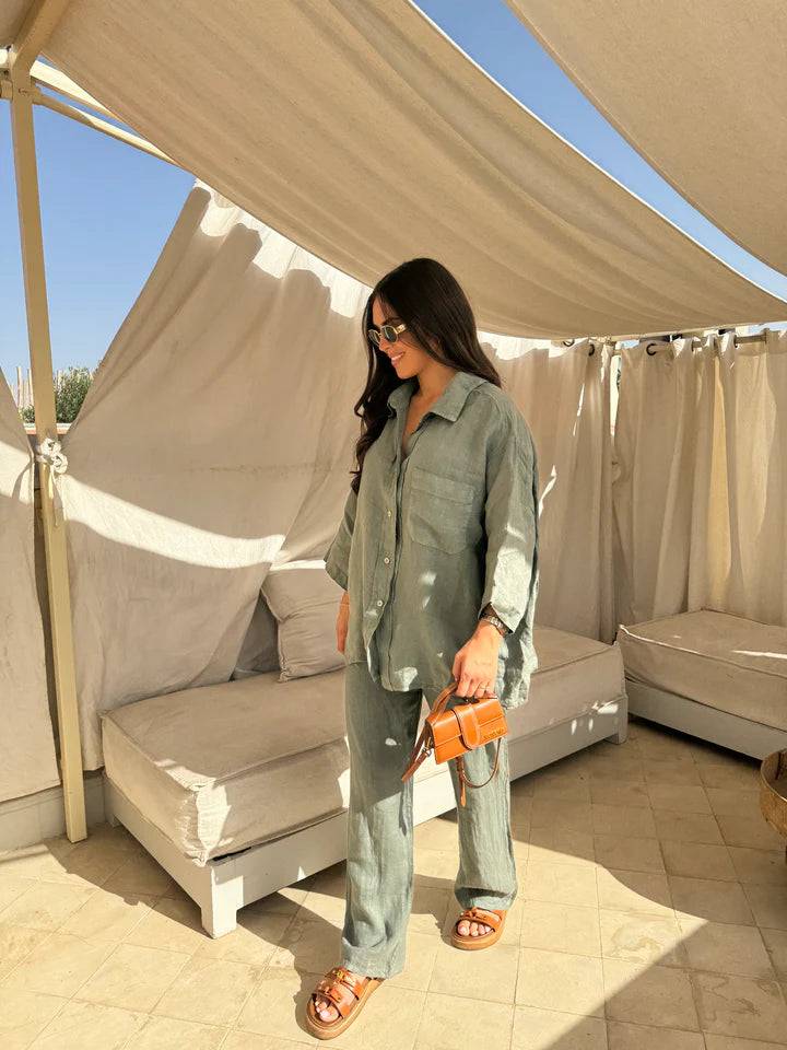 Viviana two-piece linen set in sage green, featuring an oversized button-down shirt and relaxed-fit pants, worn with brown slides and a small brown handbag on a patio.