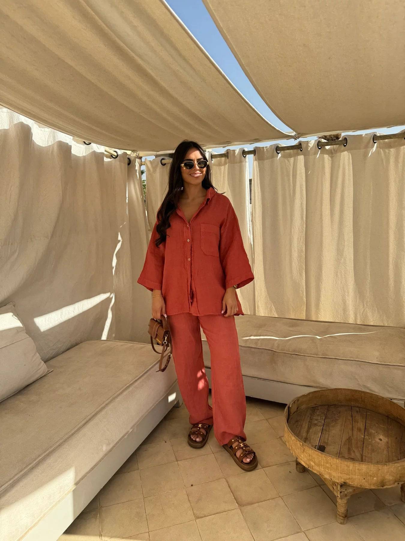 Viviana two-piece linen set in a vibrant coral-red, featuring a relaxed-fit button-down shirt with wide sleeves and matching wide-leg slacks. The model wears brown sandals and sunglasses.