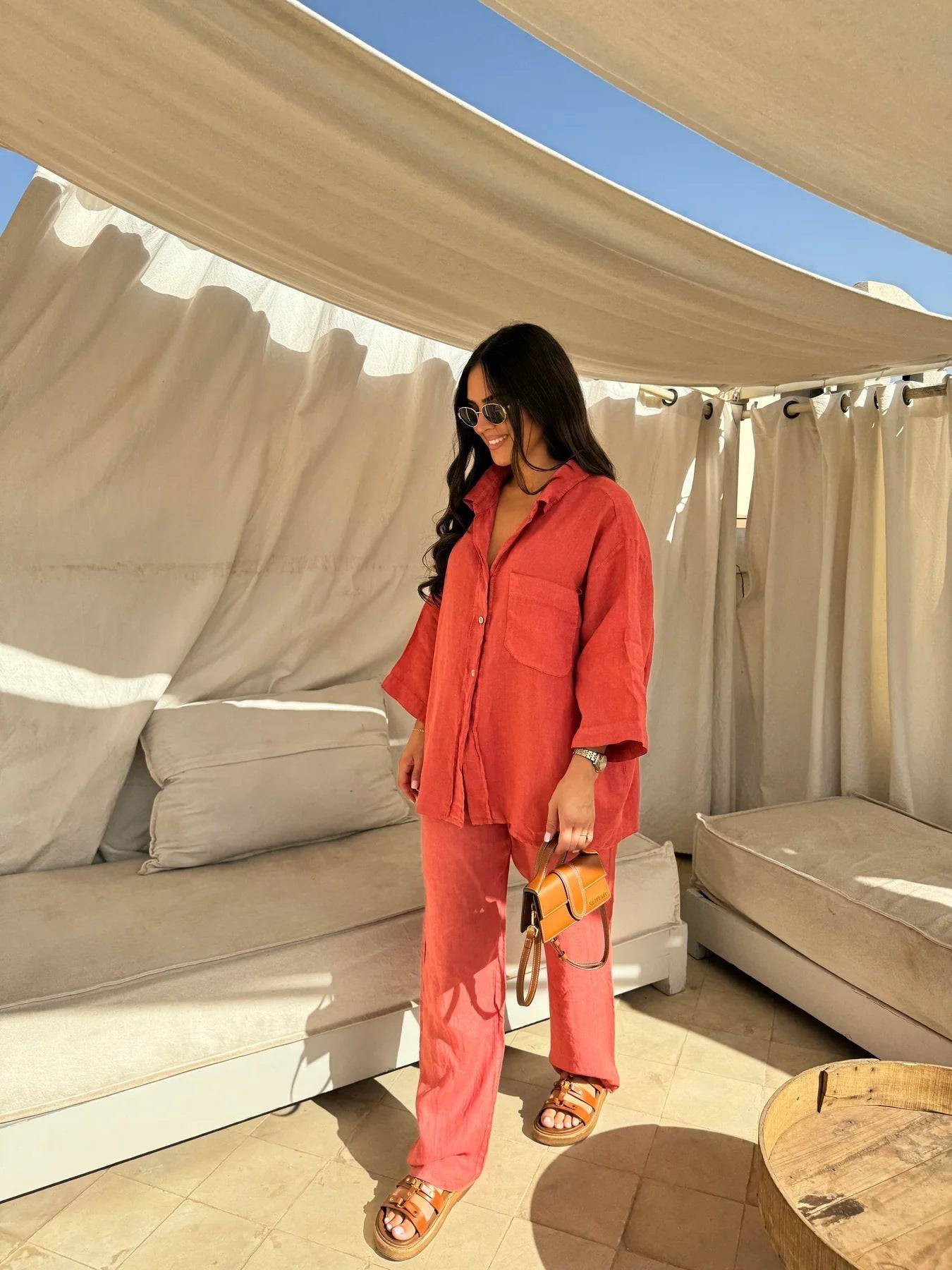 Viviana two-piece linen set in coral-red, showing the relaxed fit of the button-down shirt and comfortable wide-leg slacks. The model glances down, holding a small brown bag.