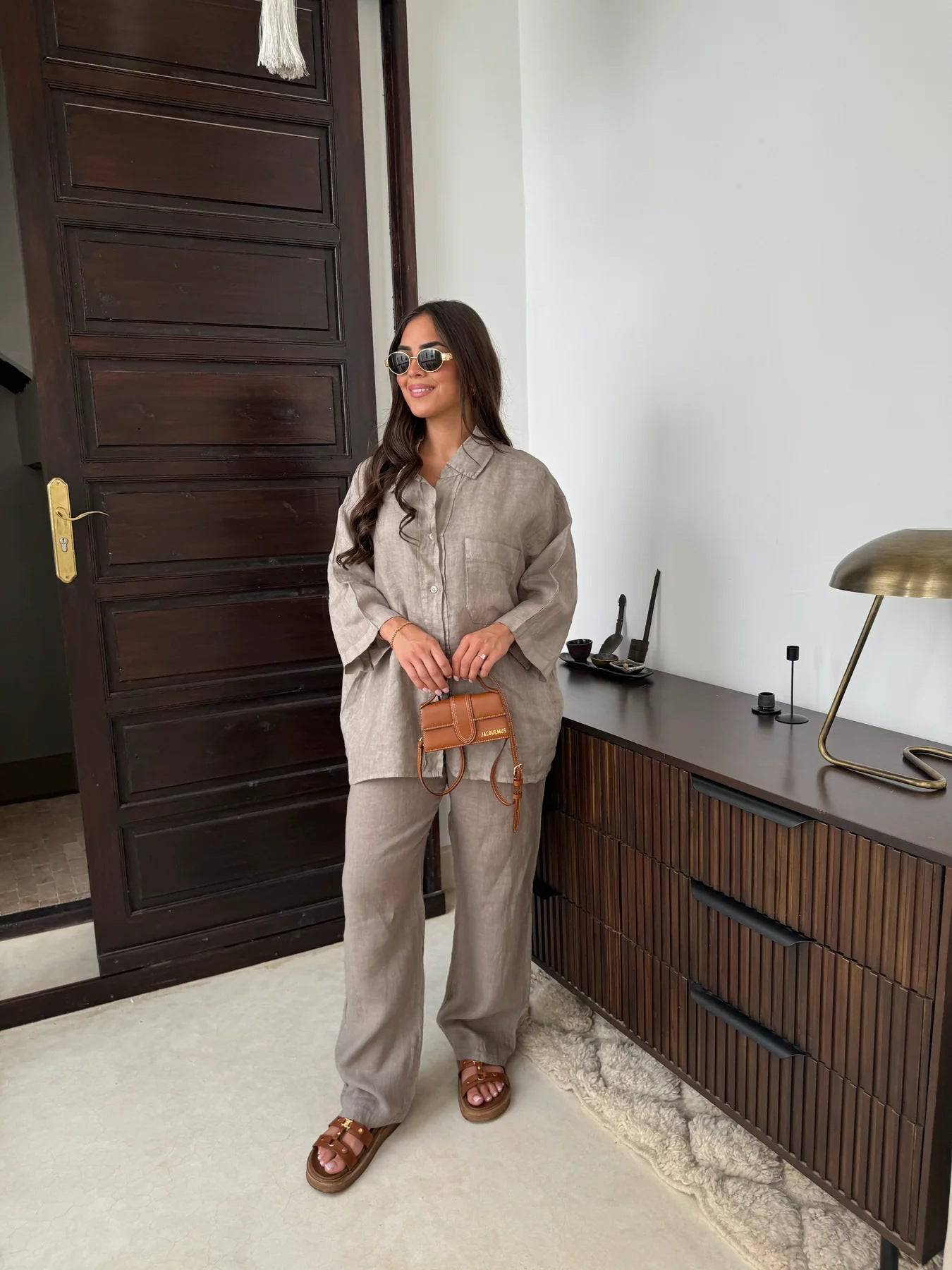 Viviana two-piece linen set in light brown, showcasing a loose-fitting shirt with a single front pocket and matching pants, paired with brown slides and a mini handbag.