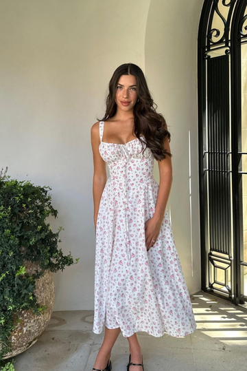 White floral midi dress with pink flower print, ruched bust, and front tie detail, worn outdoors.
