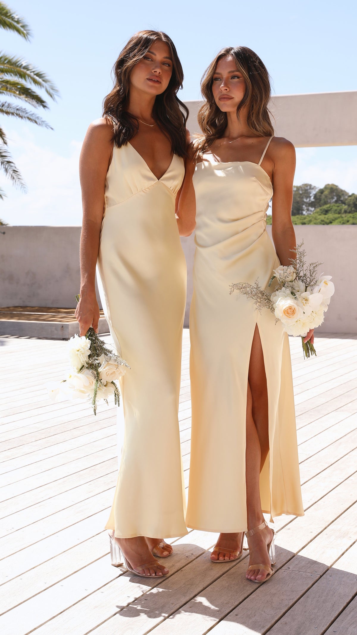 Two women standing together in pale yellow satin maxi dresses, one with a V-neck and the other a slip dress, holding floral bouquets.